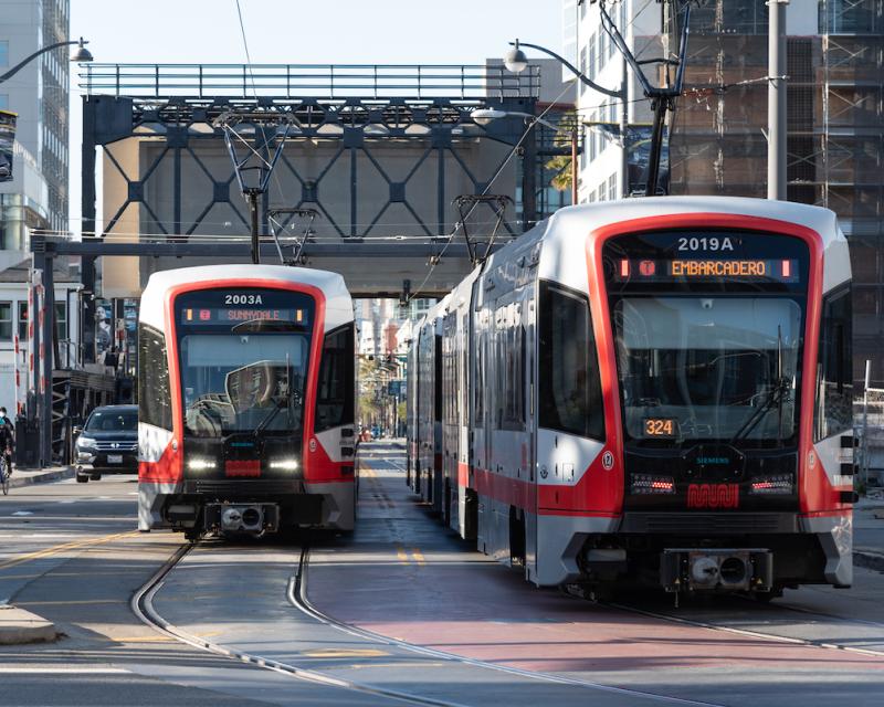 Two light rail trains pass each other on a street in San Francisco with a bridge hanging overhead in the background.