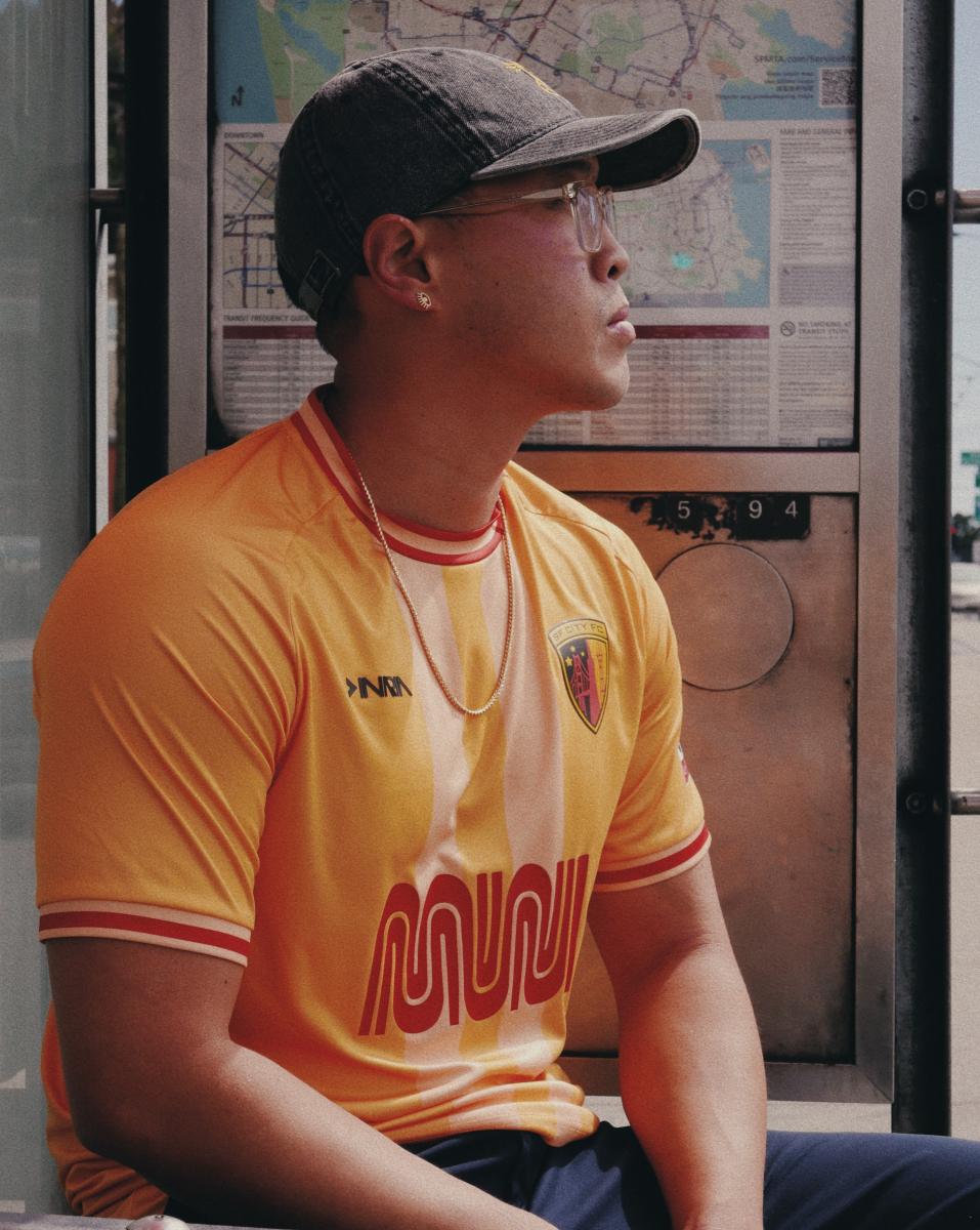 An SF City FC player wears a yellow-orange jersey with the Muni logo on it as they wait at a Muni bus shelter.