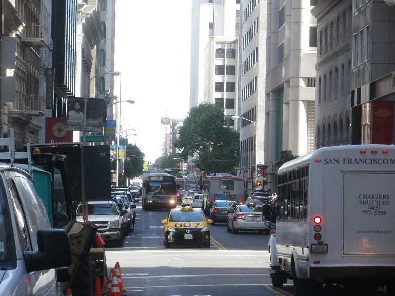 A view of Sansome Street in the Financial District. The single southbound traffic lane is used by a Muni bus and taxi, while several lanes in the opposite direction are used by  cars, buses and a shuttle van.