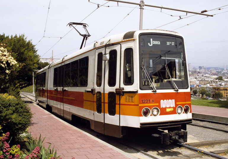 color photograph showing a Muni Boeing LRV climbing the hill in dolores park in the 1980s.  The car is stopped near the top of the hill with a view of the city skyline behind it.