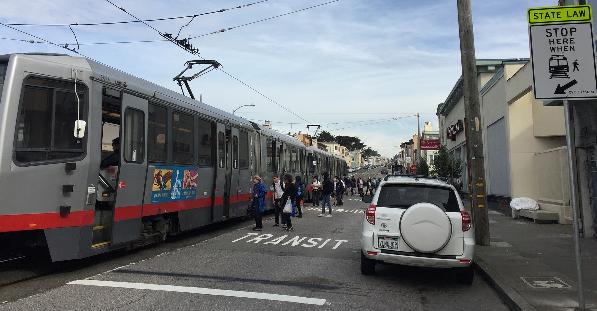 People on Taraval Street board a stopped Muni train from the roadway with pavement markings that say, "TRANSIT BOARDING ZONE" and a posted sign that says, "STATE LAW - STOP HERE WHEN" along with an image of a train and passenger.