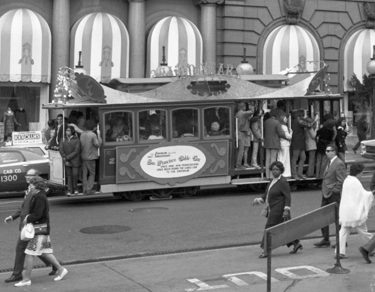 Black and white photo from august 1973, a side view of cable car 23 on Powell Street in front of the St. FRancis Hotel.  The car is covered with decorative, sparkling panels to celebrate the 100th anniversary of the cable car system.