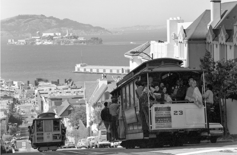 Black and white photograph taken in august of 1973 showing Cable Car 23 descending the Hyde Street hill from Chestnut Street. San Francisco Bay and Alcatraz island can be seen in the background.