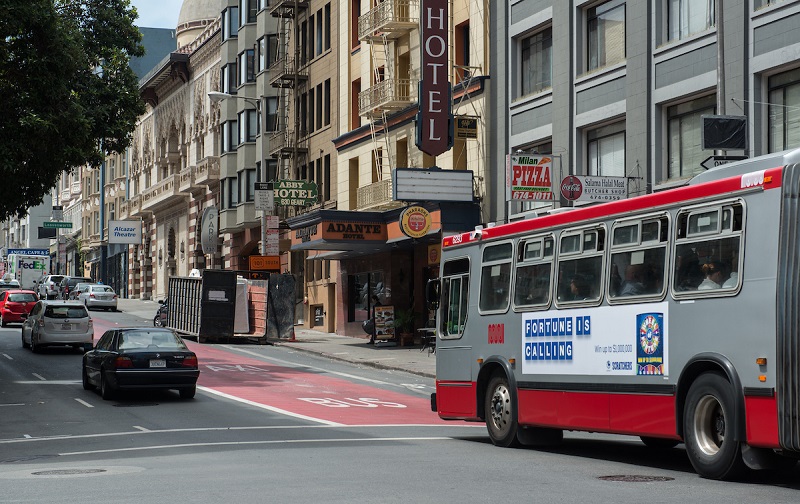 A Muni bus travels down a red transit-only lane on downtown Geary Street as cars travel in the adjacent traffic lanes.