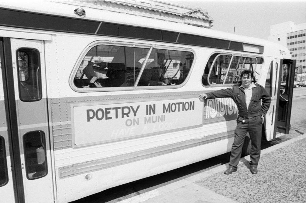 person wearing muni uniform standing next to bus with ad reading "poetry in motion on Muni, have a look!" on side of bus