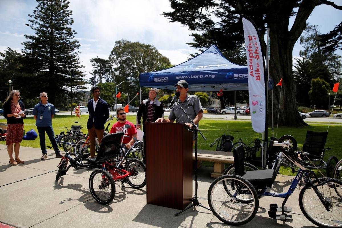 (From left: Annette Williams of SFMTA, Lucas Tobin of SF Recreation and Parks, Neal Patel of Lyft, Kurt and Finn who are BORP participants, and Greg Milano of BORP stand in Golden Gate Park on a sunny day surrounded by different kinds of adaptive bikes. Greg delivers remarks at a podium.)