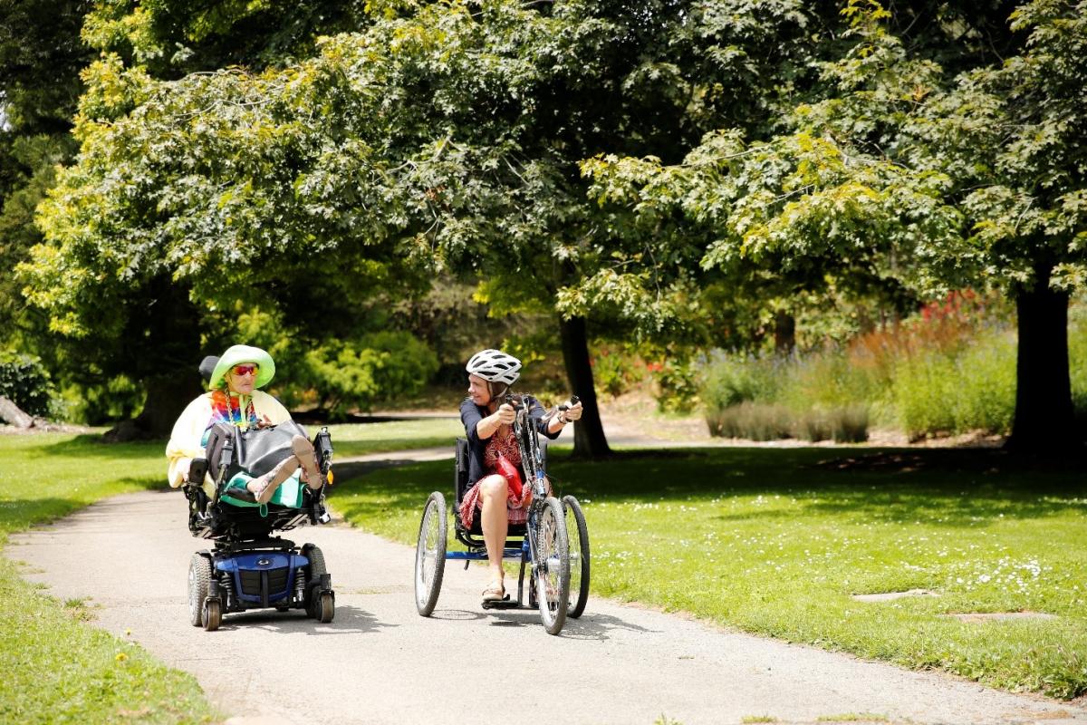 (Image: Sam-Alicia, a member of the Paratransit Coordinating Council at SFMTA, and Annette Williams of SFMTA, move side-by-side down a paved pathway surrounded by trees and green grass on a sunny day. Sam-Alicia uses her powered scooter, while Annette uses the upright hand-pedal cycle.)