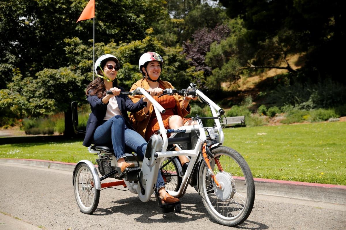 (Image: Erin McAuliff of SFMTA and Natasha Opfell of SF Paratransit sit side-by-side on the white, side-by-side tandem bike, moving down a paved road on a sunny day.)