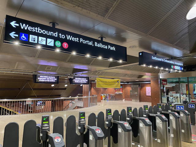 Photo of subway station fare gates with new overhead wayfinding signs depicting east, west directions and destinations for trains, and modes of accessing the platform such as elevators, stairs and escalators