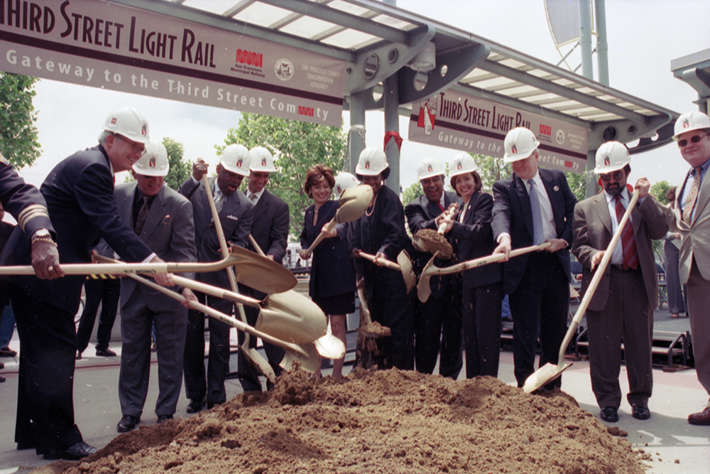 People in business attire and hard hats smiling with shovels in hand over a large pile of dirt under s station structure