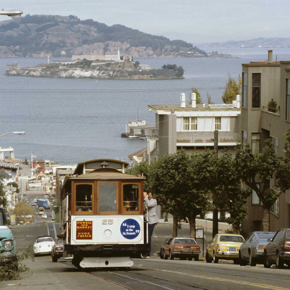 Color photo showing cable car 25 climbing hyde street with San Francisco bay and alcatraz island in background.