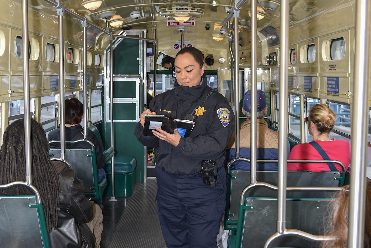 An inspector standing on a streetcar with people sitting around her. 