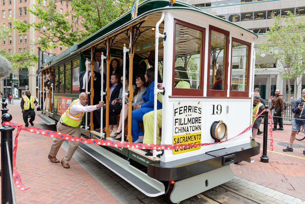 Color photo of a cable car with people riding on board and a man giving it a push start.