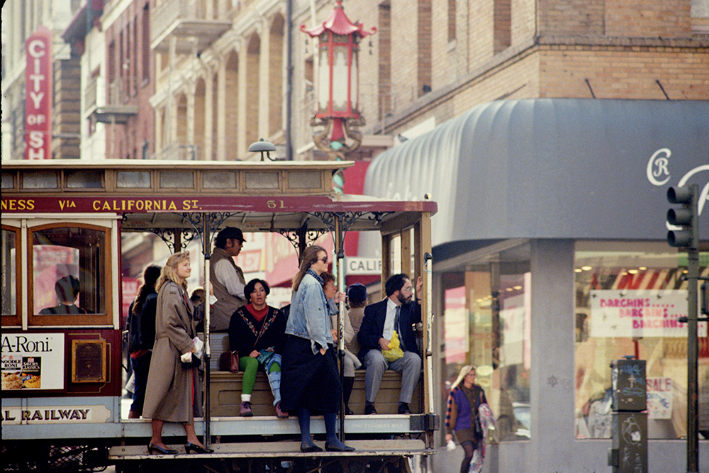 Color photo of the side of a cable car with people sitting on board and standing on the running boards.