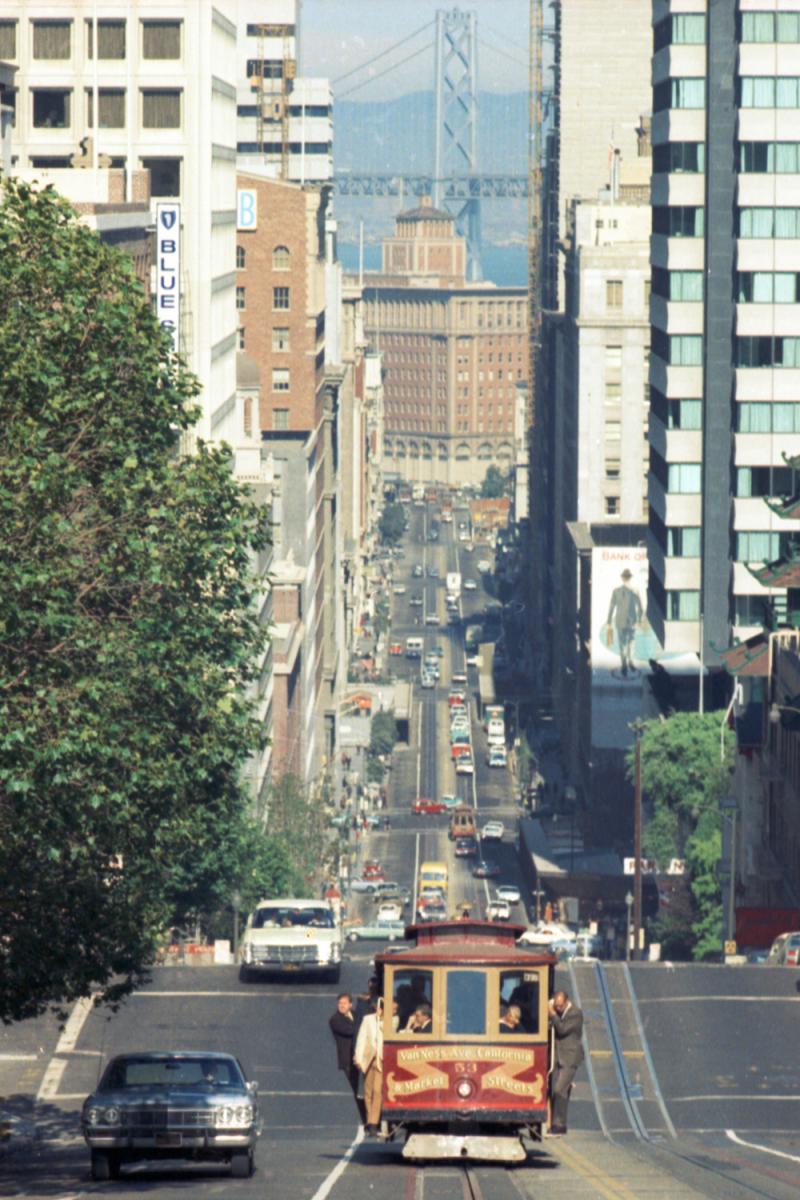 Cable Car 53 climbs up Nob Hill on California street; riders in business clothing hold as the car climbs. Bay bridge is seen in the background
