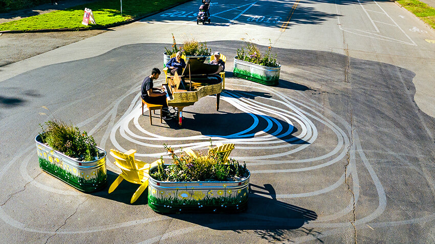 A person playing a yellow piano on a street lined by fields in a park. There are planter boxes and other artwork surrounding the piano.