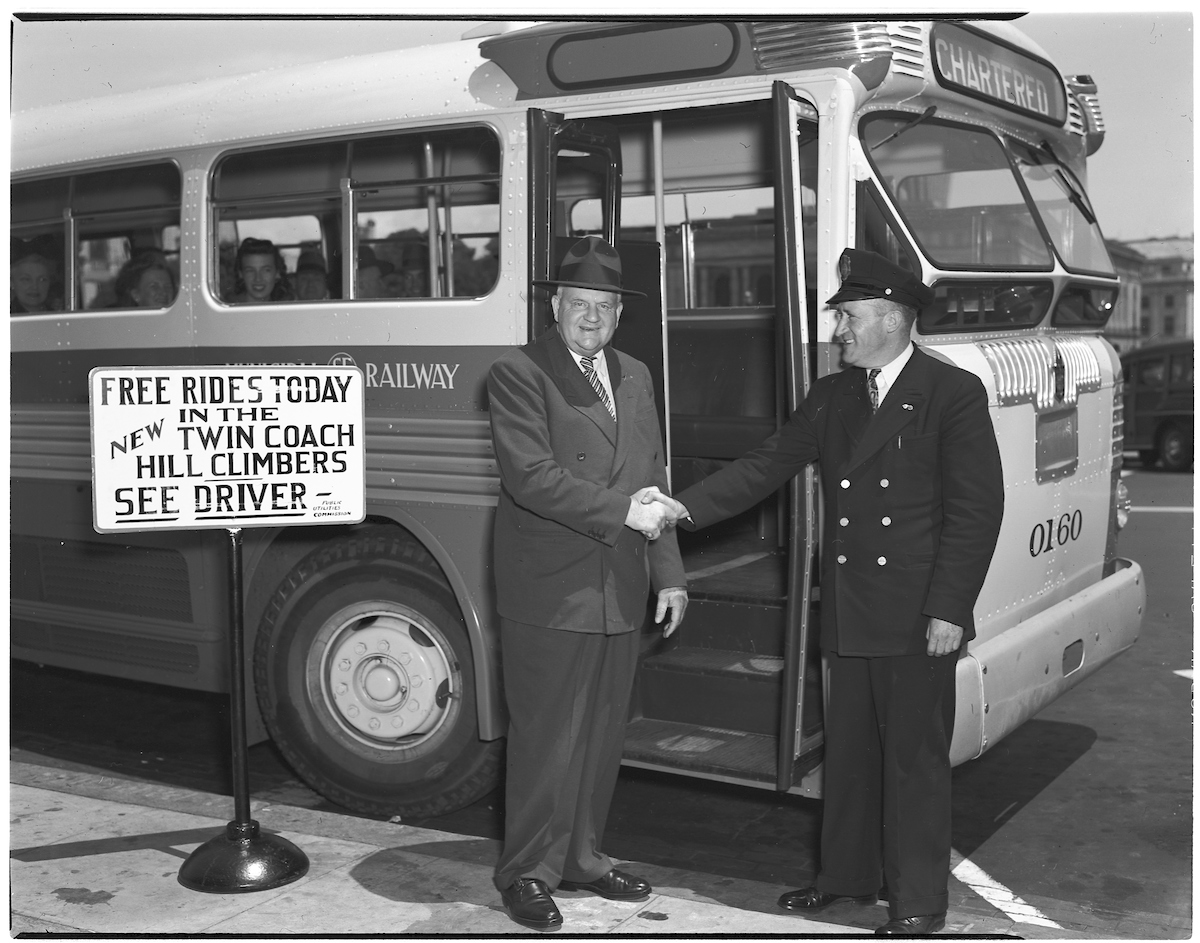 In a black and white photo, two men shake hands in front of a bus. A sign offers free bus rides for the day.