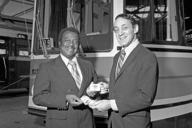 Muni General Manager Curtis Green and Supervisor Harvey Milk exchange paper money for a Muni fast pass with a light rail vehicle in the background.