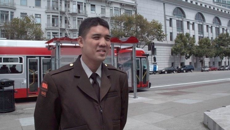 Man in a brown Muni uniform stands on a city street in front of a transit shelter and bus.