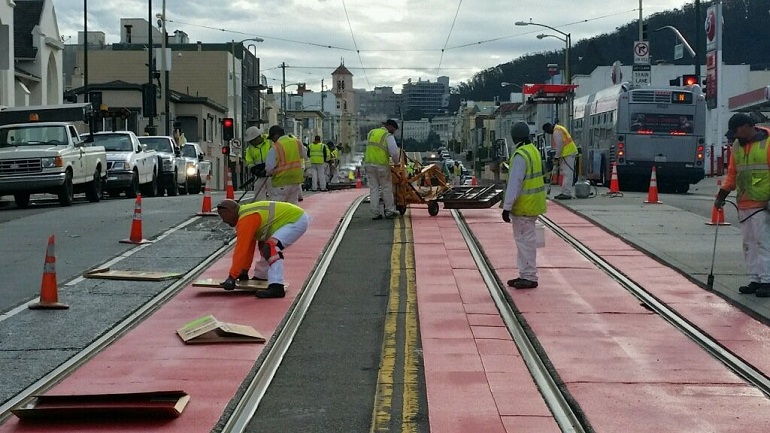 Paint crew of nine men in white pants and shirts with yellow safety vests works in the middle of Judah Street looking east to install the red thermoplastic tiles around the center trackway.