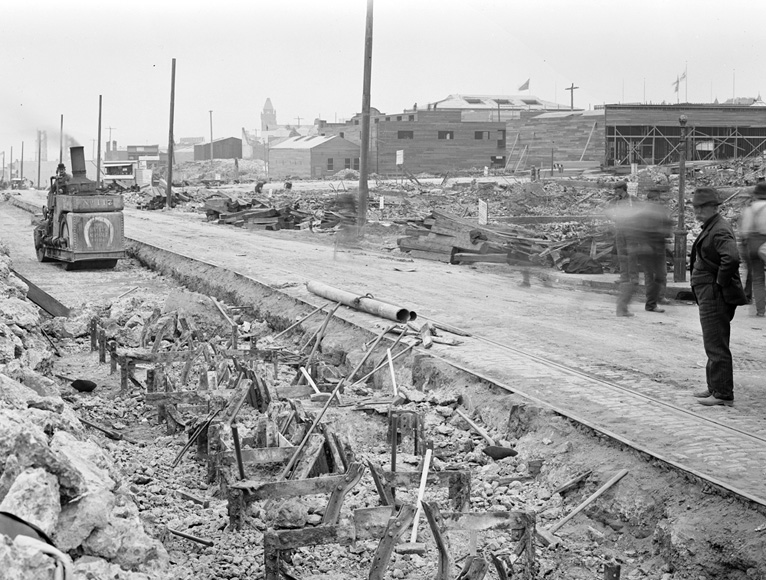 An image taken on July 20, 1906 shows a trench of metal structures from cable car tracks. Men appear to be at work to remove them, one of them driving a machine approaching the structures.