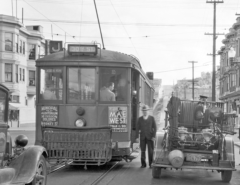 black and white poto taken in 1934 on Church and 17th Streets.  A streetcar o the J line is stopped next to a fire truck and a man is standing between the fire truck and streetcar facing the camera.  The streetcar has a movie poster advertisement on the back reading "mae west"