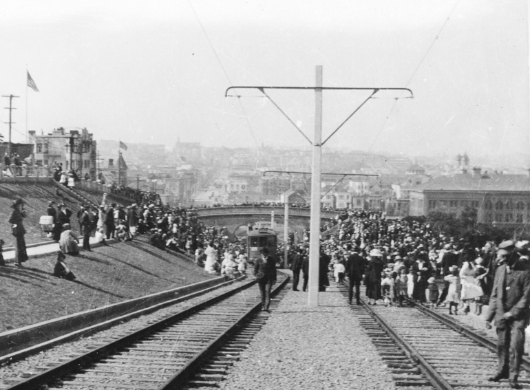 black and whit photo taken August 11, 1917 showing the opening ceremonies of the J Line.  A crowd of people swarms the tracks and pedestrian bridge in Dolores Park as streetcars make their way up the hill.