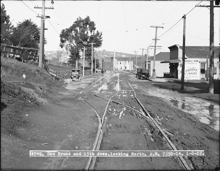 Black and white photograph showing intersection of Bayshore Boulevard and Oakdale Avenue on January 8, 1915. Streetcar tracks receed into the distance away from the camera. to the left is a hillside covered with grass, trees, and some wooden fencing.  To the right is a small, flat-fronted building which houses the "Oakdale Bar", today this houses the Old Clamhouse restaurant.