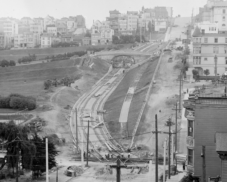 black and white photo taken in June 1916, looking south on church street from 17th at the construction of the J Line through Dolores Park.  The trackway is cut into the hillside in a serpentine fashion and workers are scattered about along the route digging and installing tracks.