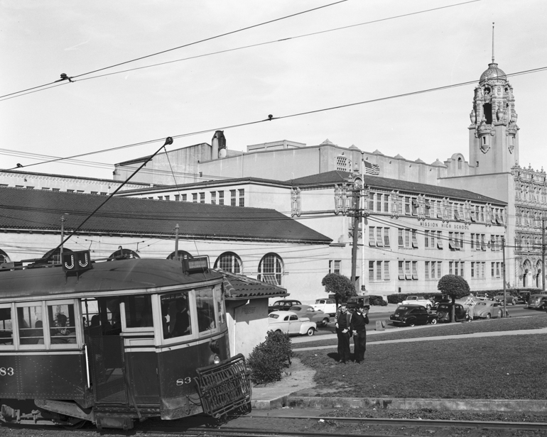 black and white photo showing a streetcar passing Mission High school in dolores park.  Two teenaged boys are standing to the right of the streetcar in the grass.