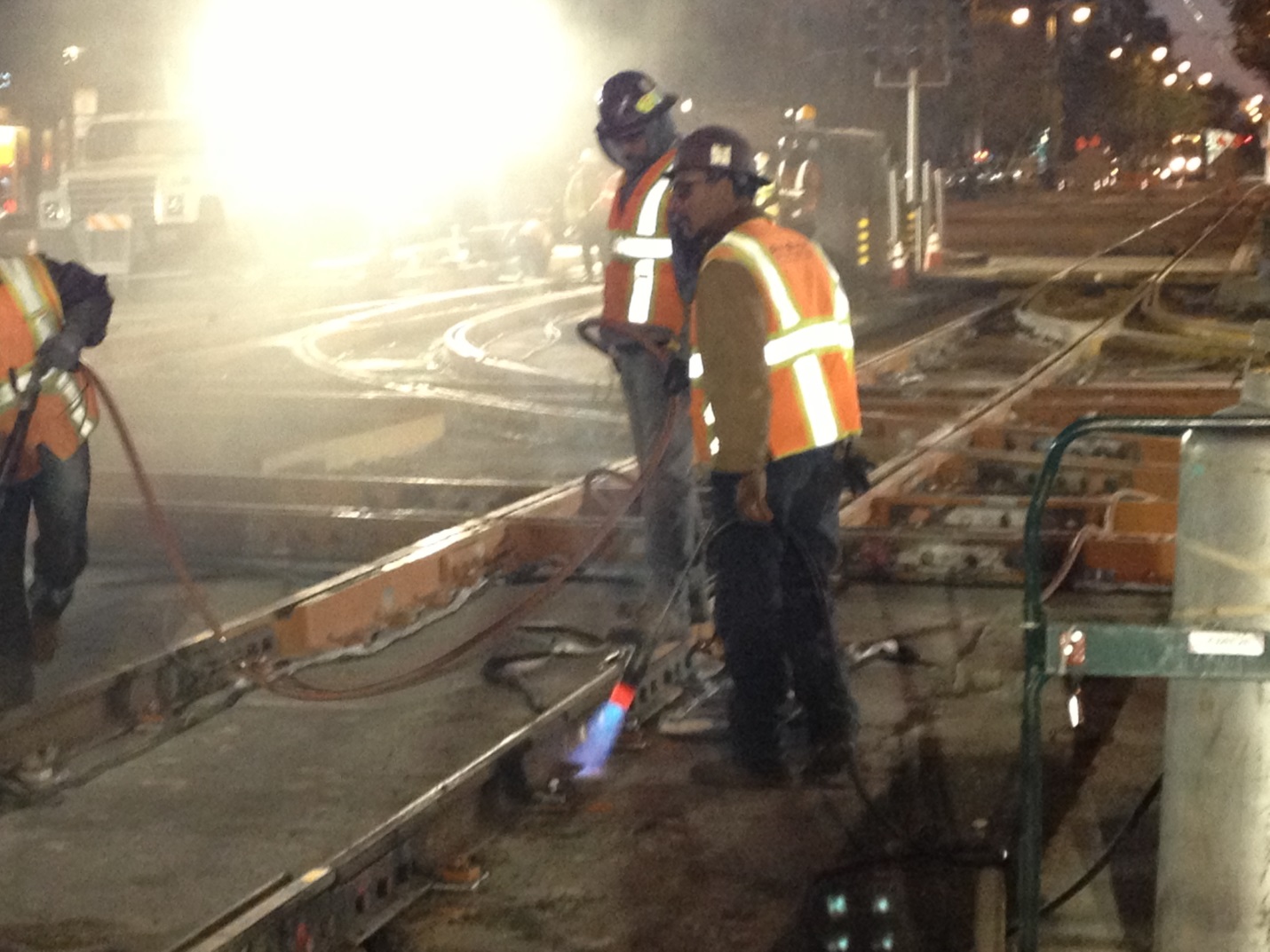 Crew members in safety vest and hard hats stand next to tracks being installed at 4th and King at night. Welding gear and safety strips are glowing 