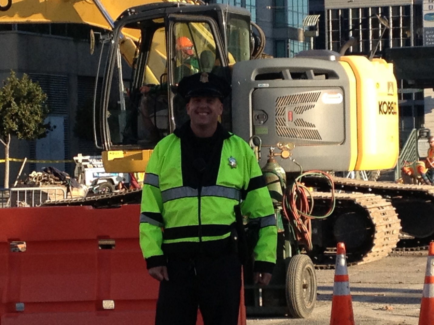 A police officer wearing a reflective safety jacket stands in front of construction equipment.