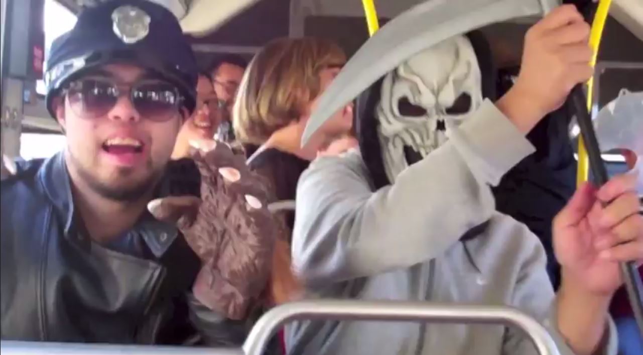 People in costume sitting on a Muni bus: man on left wears a police hat, sunglasses and werewolf gloves; person on right wears a skeleton mask, great sweatshirt and is carrying a scylthe. People in background are singing.