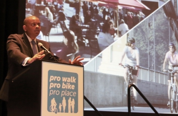 Transportation Secretary Foxx stands at a lecturn labeled "Pro Walk, Pro Bike, Pro Place." Photographs of bicyclissts and people at a cafe are projected behind him.