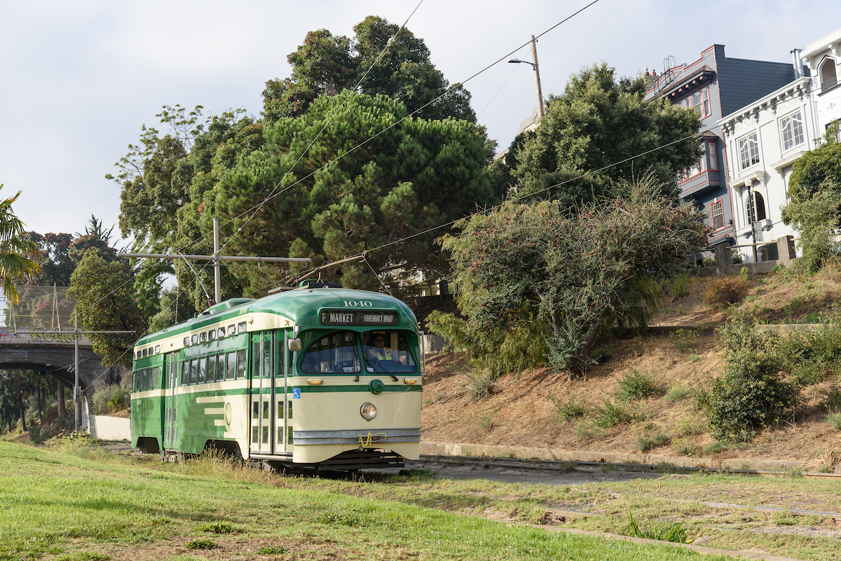 Historic streetcar painted cream and green moves through Dolores Park with a bridge in the background and grass in the foreground.