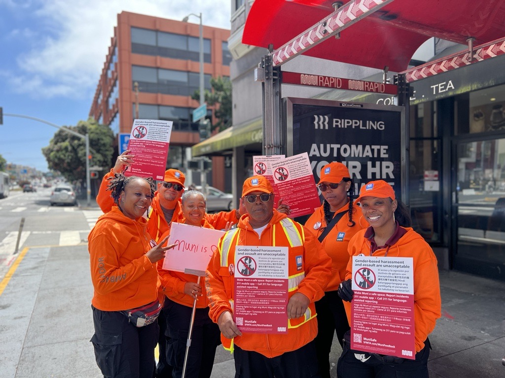 MuniSafe ambassadors and staff smile at a bus stop, holding signs sharing how to report harassment. They wear bright orange hoodies.