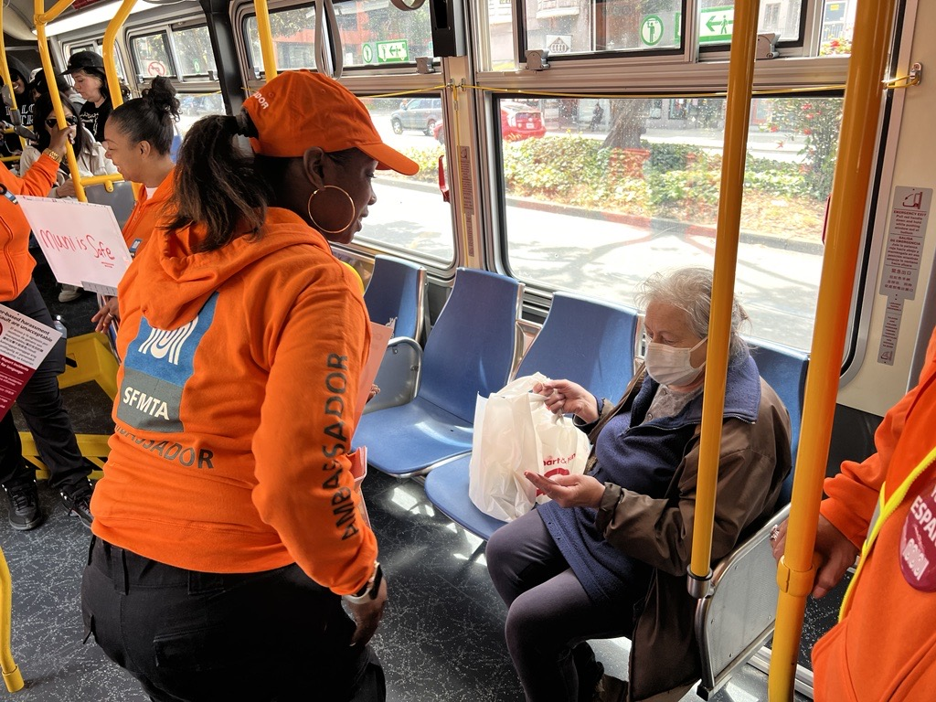 A MuniSafe ambassador wearing a bright orange hoodie and hat chats with a rider on a Muni bus about MuniSafe.