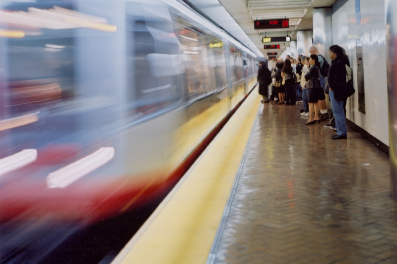 A view of a Muni Metro platform in 1999 with a train in motion on the left while passengers stand on the platform on the right.