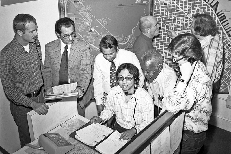 A group of Muni employees from the Muni Public Service Bureau cluster around a man with a headset inside the Muni Offices in 1977.
