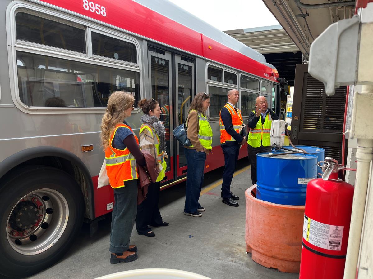 several people in safety vests stand near a bus and look at examine and discuss nearby repair equipment