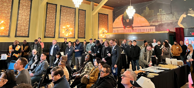 dozens of people are shown sitting or standing in a spacious and elegantly decorated room. Most are looking off camera in the same direction while a few others are looing at information on a table. 