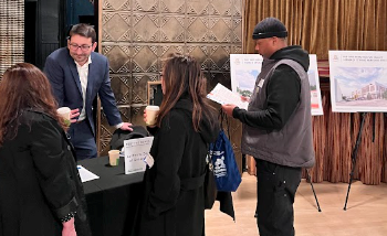  person stands behind a table talking to a few other people gathered nearby. Display boards on easels are visible. Architectural elements and wall treatments are seen in the background.