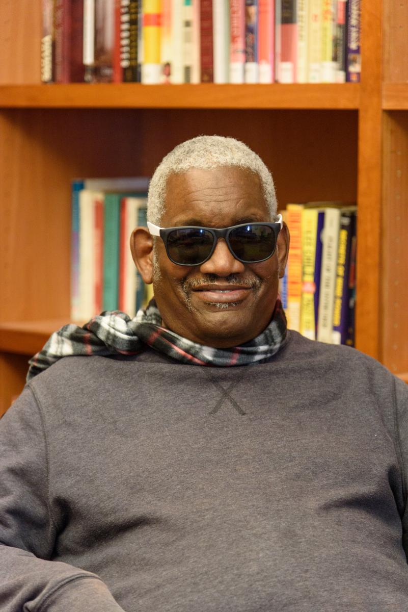 Dennis, an elderly Black man is pictured seated and smiling, in front of bookshelves. He is wearing a sweater, a scarf, and sunglasses.