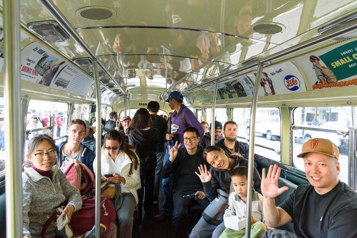 A group of people sit in a vintage bus. A few of them wave to the camera.