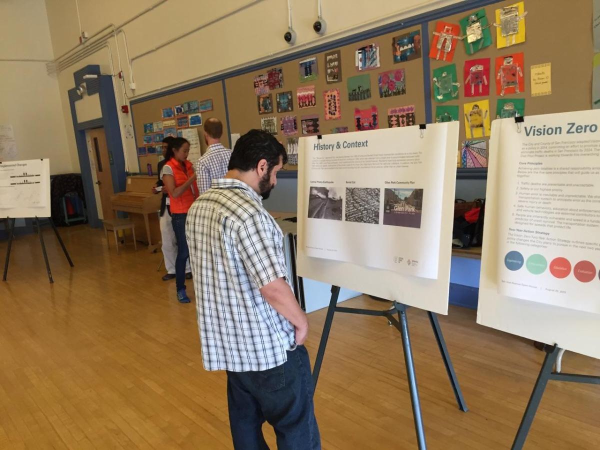 Man viewing presentation board labeled "History & Context" at community open house event in school gymnasium.