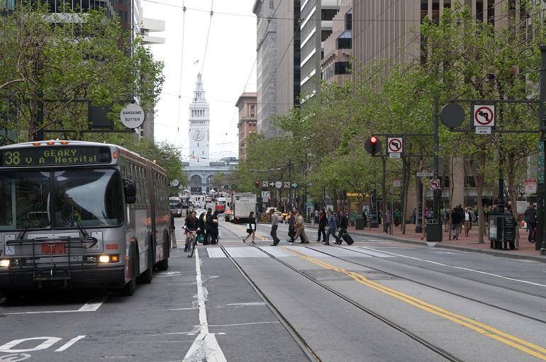 Market Street scene with Muni bus, streetcar tracks, pedestrians, and Ferry Building in background.