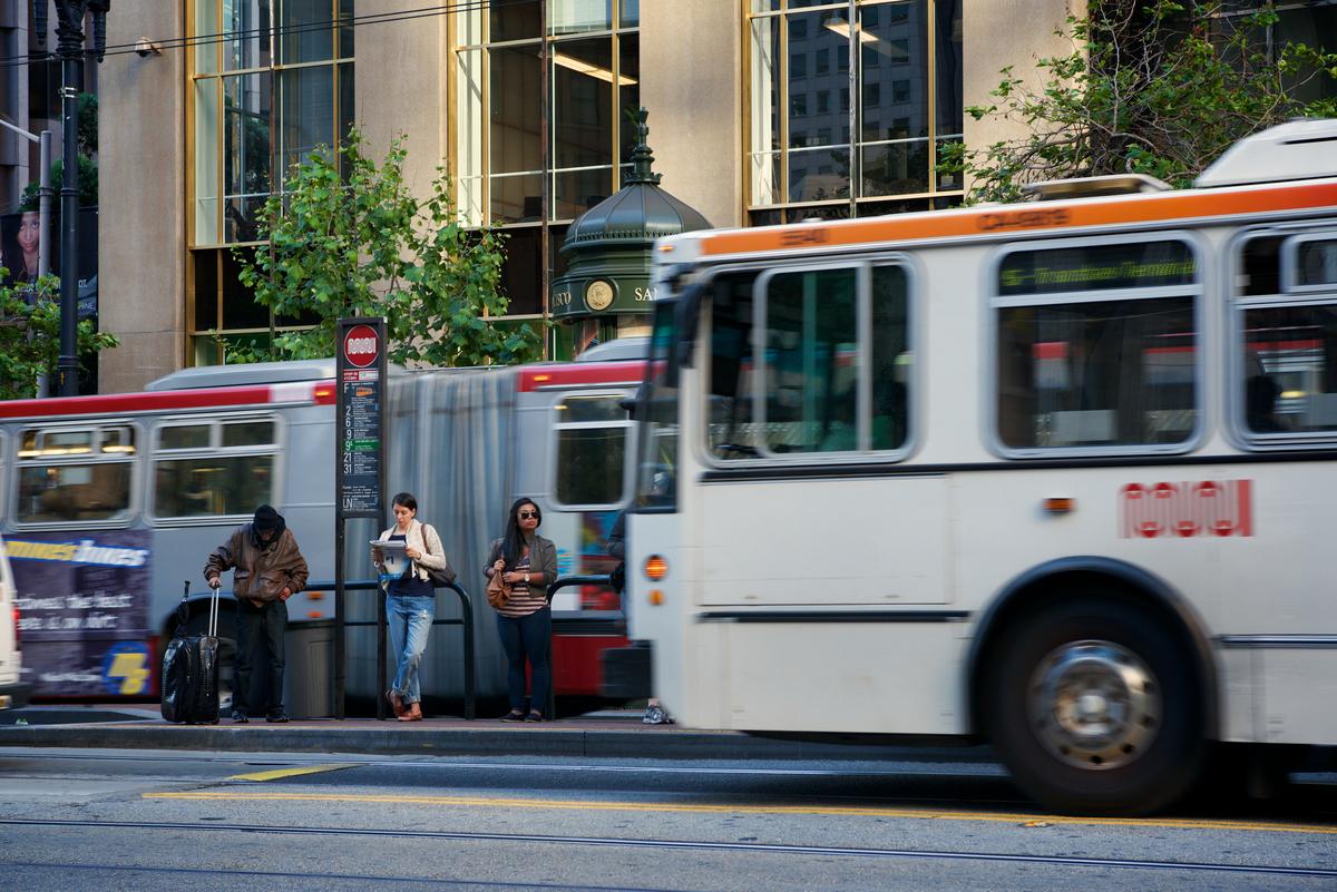 Three passengers wait at a bus stop as a Muni bus approaches on a city street.