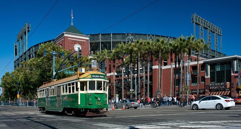 Green and cream streetcar passing AT&T Park with palm trees and brick building under blue sky.