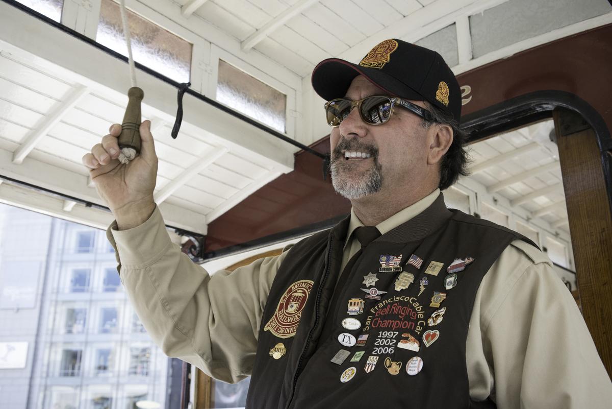 Man in cable car uniform and cap rings bell overhead during competition.