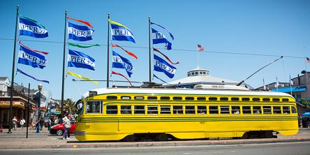 Yellow streetcar stopped at Pier 39 with blue flags and San Francisco waterfront in background